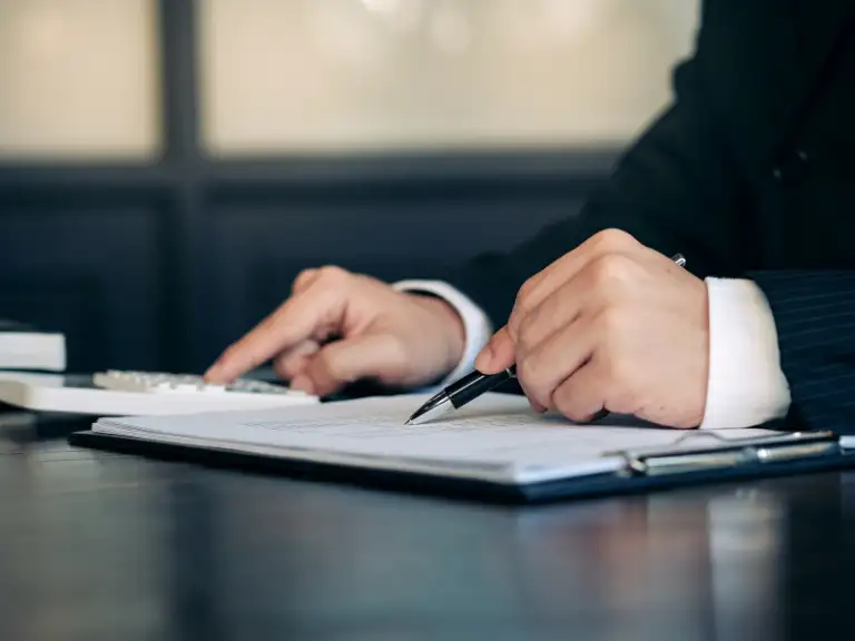 Profissional com terno e camisa branca usando uma calculadora e caneta sobre documentos em uma prancheta